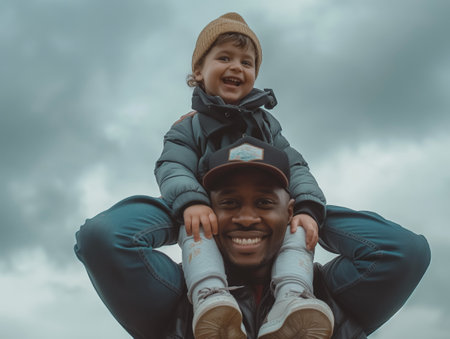 A smiling child sits atop their fathers shoulders against a cloudy sky background, depicting family happiness and bonding.の素材