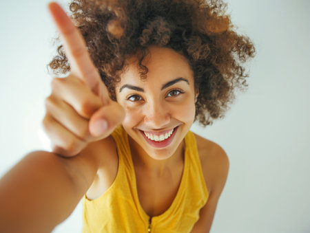 A smiling young woman with curly hair pointing towards the camera on a light background, conveying a friendly and engaging atmosphere.の素材
