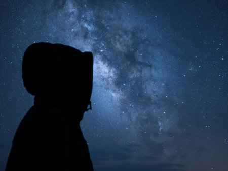 Silhouette of a person in a hooded jacket looking up at the Milky Way galaxy in a clear night sky.の素材