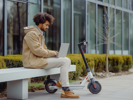 A man in a beige jacket and white pants sits on a bench, working on a laptop beside an electric scooter.の素材