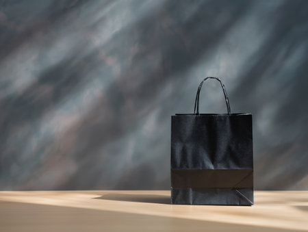 A black paper bag with handles sits on a wooden surface against a blurred, multicolored backgroundの素材