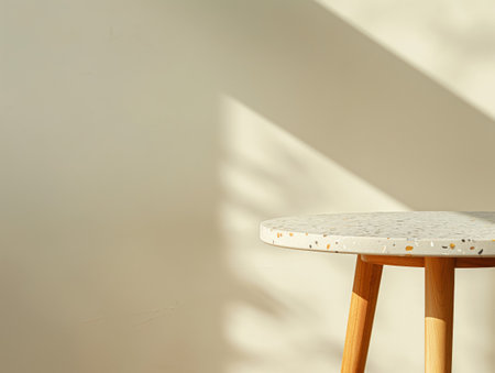 A round table with a terrazzo top and wooden legs is bathed in soft sunlight, casting shadows on a plain wall.の素材