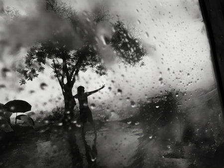 A silhouette of a person stands with an outstretched arm under a tree in the rain, viewed through a rain-soaked window.の素材