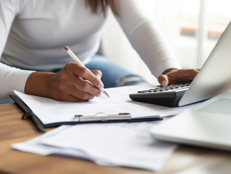 A person writes on a document with a pen while using a calculator, with a laptop nearby on a desk.の素材