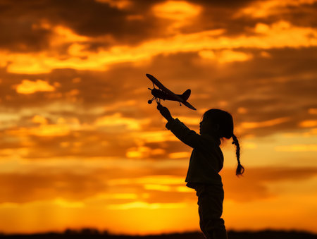 Silhouette of a child with a toy airplane, set against a vibrant orange sunset sky with scattered clouds.の素材