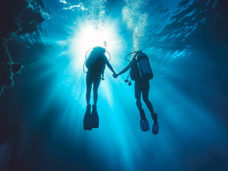 Two scuba divers hold hands while swimming underwater, illuminated by sunlight filtering through the waterの素材