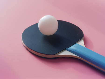 A close-up of a ping pong paddle with a blue handle and a white ball resting on its black rubber surface, set against a pink backgroundの素材