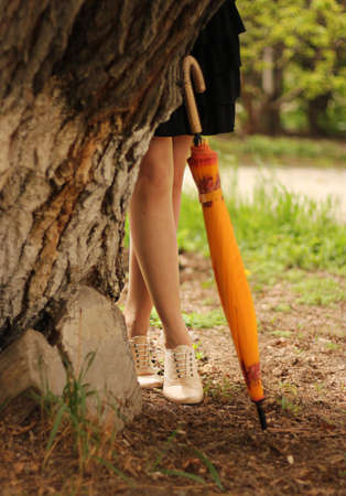 girl is standing behind the tree with only legs visible and an umbrella is put by that same treeの写真素材