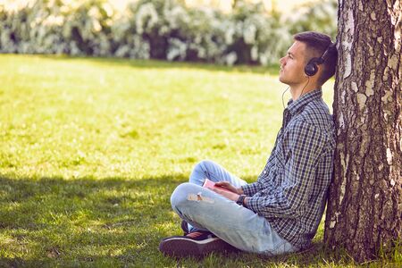 Music, technology, people and leisure concept - close up of teenage boy in headphones with book listening to music online at parkの写真素材