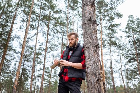 Man with beard taking rest in forest after walk, is drinking hot coffee