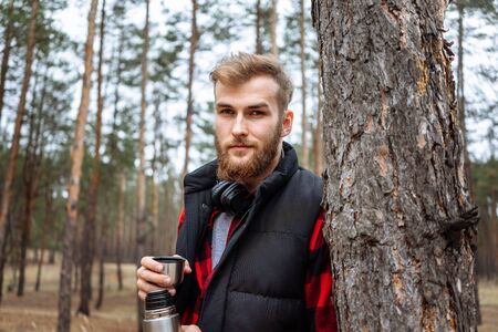 Man with beard taking rest in forest after walk, is drinking hot coffee