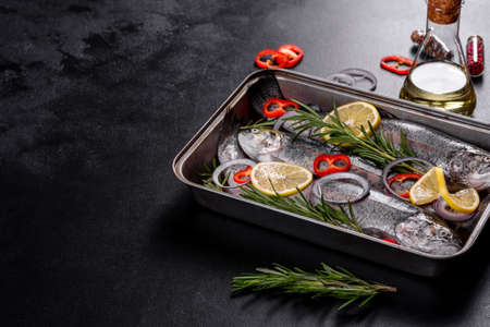 Raw trout lie on a black stone chopping board on a dark concrete table. Nearby are: tomato,...