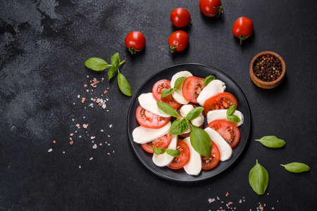 Italian caprese salad with sliced tomatoes, mozzarella cheese, basil, olive oil. Served in vintage black plate on a dark concrete backgroundの写真素材