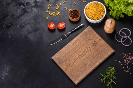 A wooden cutting board with a kitchen knife with spices and herbs on a dark concrete background....
