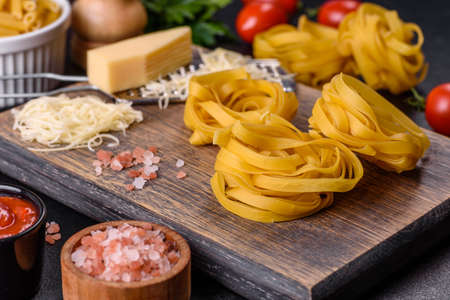 Raw tagliatelle paste with grated cheese, cherry tomatoes, spices and herbs on a wooden cutting board against a dark concrete backgroundの写真素材