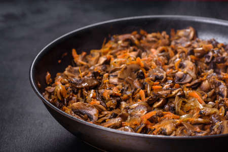 Fried champignons with carrots, onions and spices in a pan against a dark concrete background. Vegetarian cuisineの写真素材