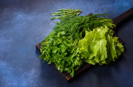 Salad, parsley and dill on a dark cutting board against a blue concrete background. Cooking a vegetarian dish with fresh greensの写真素材