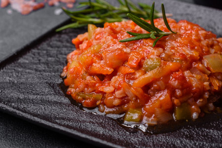 Boiled rice with peppers, spices, herbs and tomato sauce on a black slate plate on a gray concrete...
