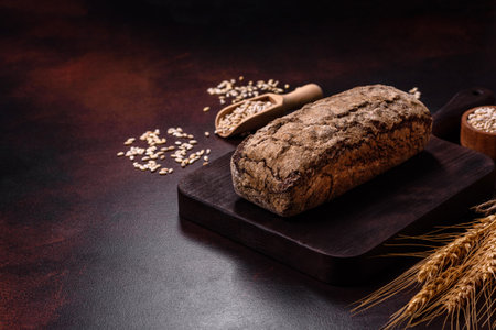 A loaf of brown bread with grains of cereals on a wooden cutting board on a dark concrete background