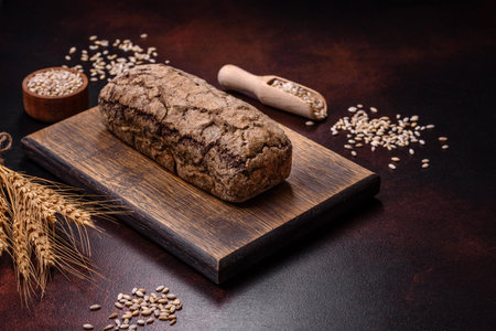 A loaf of brown bread with grains of cereals on a wooden cutting board on a dark concrete background