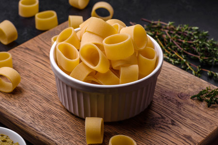 Raw uncooked pasta in a white ceramic bowl with spices and herbs on a dark concrete background