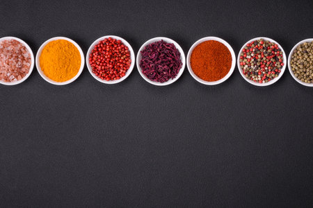 Composition consisting of variations of spices in white bowls and metal spoons on a dark concrete...