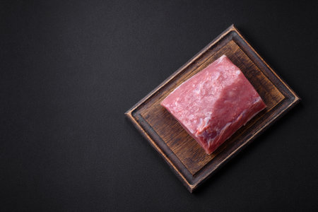 A piece of raw fresh pork on a wooden cutting board with spices and herbs on a dark concrete backgroundの写真素材