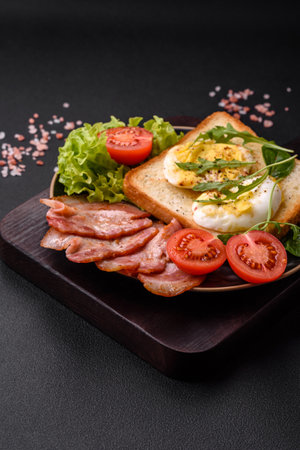 Delicious healthy lunch consisting of bacon, toast, eggs, tomatoes, lettuce and greens on a black plate on a concrete backgroundの写真素材