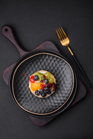 Delicious fresh tart with blueberries, cherries, grapes on a black ceramic plate on a dark concrete backgroundの写真素材
