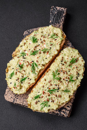 Grilled toast with avocado paste, eggs, cream cheese, salt, spices and herbs on a textured concrete tableの写真素材