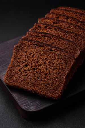 Delicious fresh brown bread with grains and seeds sliced on a wooden cutting board on a dark concrete backgroundの写真素材