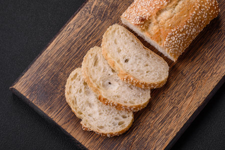 Crispy wheat flour baguette with sesame seeds on a wooden cutting board on a dark concrete backgroundの写真素材