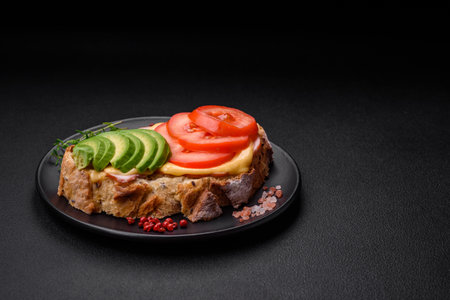 Delicious nutritious grilled toast with ham, cheese, tomatoes and avocado with salt, spices and herbs on a dark concrete backgroundの写真素材