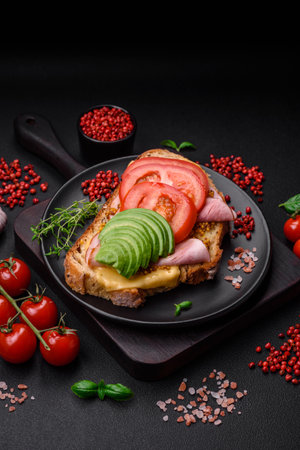 Delicious nutritious grilled toast with ham, cheese, tomatoes and avocado with salt, spices and herbs on a dark concrete backgroundの写真素材