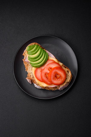 Delicious nutritious grilled toast with ham, cheese, tomatoes and avocado with salt, spices and herbs on a dark concrete backgroundの写真素材