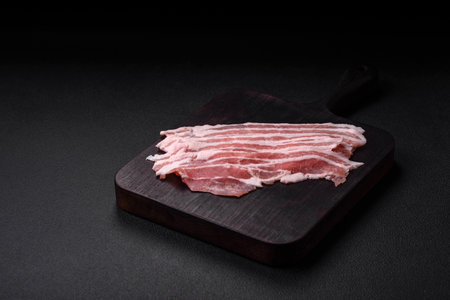 Fresh raw bacon cut into slices with salt, spices and herbs on a dark textured concrete background