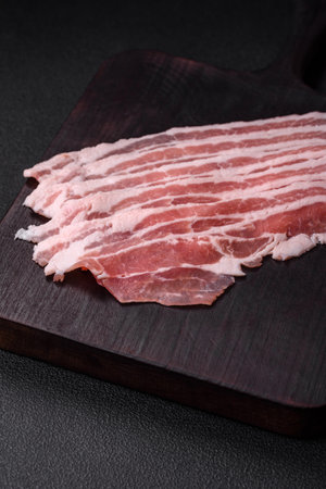 Fresh raw bacon cut into slices with salt, spices and herbs on a dark textured concrete background