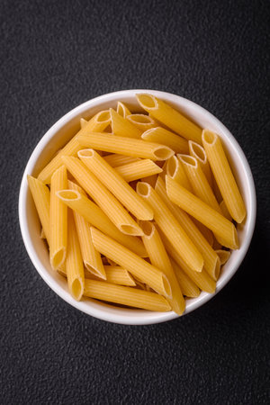 Raw penne pasta from whole grain wheat varieties with salt and spices on a dark concrete background