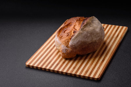 A loaf of brown bread with grains of cereals on a wooden cutting board on a dark concrete background
