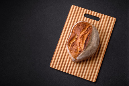 A loaf of brown bread with grains of cereals on a wooden cutting board on a dark concrete background