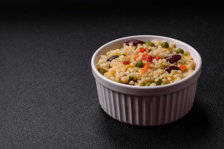 Boiled wheat bulgur porridge with vegetables peppers, peas, corn on a dark concrete backgroundの写真素材