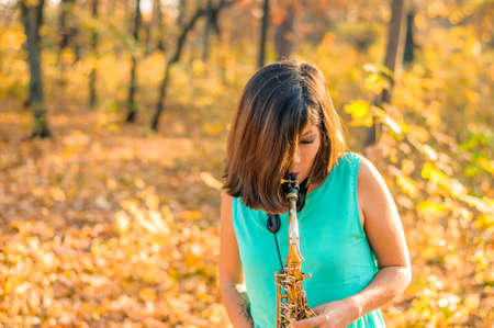 Young brunette girl with hair covering her face plays the saxophone, standing in blue clothes in a yellow autumn parkの写真素材