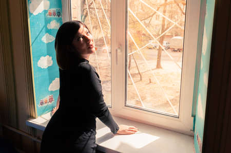 unusual beautiful young girl with black hair in a long tight dark dress stands by the window,...