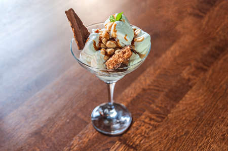 Dessert of white ice cream, peanuts and a chocolate bar with a mint leaf in a glass bowl on a wooden tableの写真素材