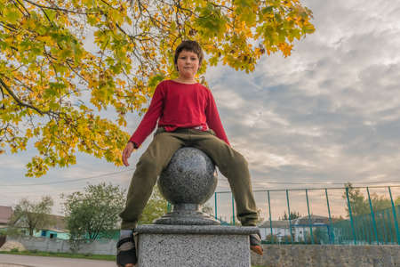 funny young boy in casual clothes sits on a decorative fence with marble ball near an autumnの写真素材