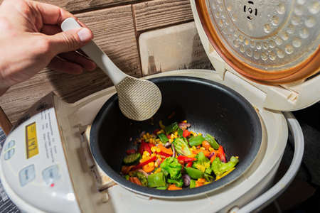 mixed set of different vegetarian vegetables in a white multicooker is stirred with a kitchen...