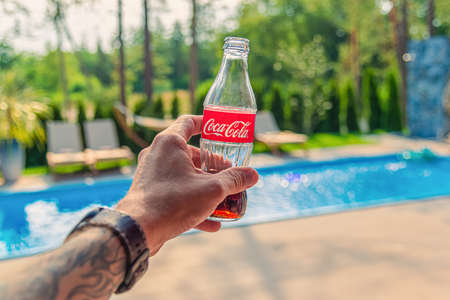Man hand with a tattoo holds a glass bottle with coca-cola on the background of the pool and trees on a sunny dayのeditorial素材