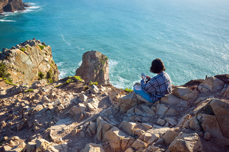 SINTRA, PORTUGAL - FEBRUARY 11, 2019: Young woman sitting is using smartphone on mountain cliff edge on ocean sunny natural background. Traveler having social communication technology.のeditorial素材
