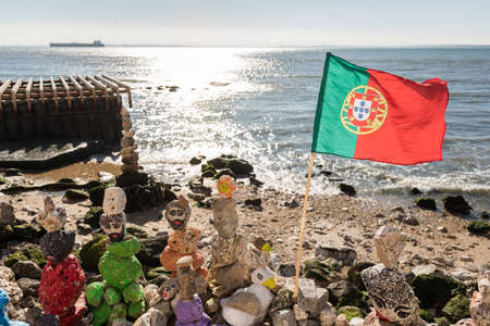 Lisbon, Portugal - February 12, 2019: Landscape view across sand beach on shore with flag, stone people structures and calm ocean beyond at the harbour in sunshine light of day.のeditorial素材