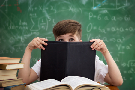 Funny schoolboy hiding behind book sitting in a classroom over a blackboardの写真素材